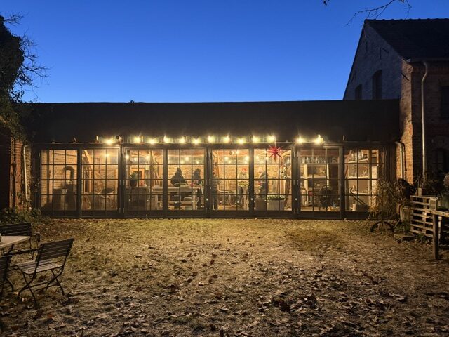 Die Wagenremise am Hof Eversmann bei Abenddämmerung — Stahlglasfenster mit warmem Licht und Lichterketten